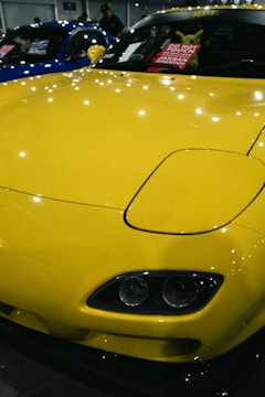 A brightly colored yellow car with a sleek and polished finish, parked indoors. The hood features striking headlights and the windshield displays a sign in multiple languages cautioning not to touch the vehicle. Reflections of lights on the car's surface add to the shiny aesthetic.