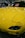 A brightly colored yellow car with a sleek and polished finish, parked indoors. The hood features striking headlights and the windshield displays a sign in multiple languages cautioning not to touch the vehicle. Reflections of lights on the car's surface add to the shiny aesthetic.
