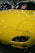 A brightly colored yellow car with a sleek and polished finish, parked indoors. The hood features striking headlights and the windshield displays a sign in multiple languages cautioning not to touch the vehicle. Reflections of lights on the car's surface add to the shiny aesthetic.