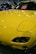 A brightly colored yellow car with a sleek and polished finish, parked indoors. The hood features striking headlights and the windshield displays a sign in multiple languages cautioning not to touch the vehicle. Reflections of lights on the car's surface add to the shiny aesthetic.
