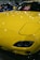 A brightly colored yellow car with a sleek and polished finish, parked indoors. The hood features striking headlights and the windshield displays a sign in multiple languages cautioning not to touch the vehicle. Reflections of lights on the car's surface add to the shiny aesthetic.