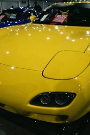 A brightly colored yellow car with a sleek and polished finish, parked indoors. The hood features striking headlights and the windshield displays a sign in multiple languages cautioning not to touch the vehicle. Reflections of lights on the car's surface add to the shiny aesthetic.