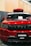 A front view of a customized red SUV with black accents. The car has a prominent grille with multiple horizontal slots, a hood scoop, and a roof-mounted cargo box. The windshield has decals, including 'STNCSG,' and a piece of paper placed on the dashboard. The background includes a wall with vertical wooden panels and part of a ceiling structure.
