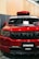 A front view of a customized red SUV with black accents. The car has a prominent grille with multiple horizontal slots, a hood scoop, and a roof-mounted cargo box. The windshield has decals, including 'STNCSG,' and a piece of paper placed on the dashboard. The background includes a wall with vertical wooden panels and part of a ceiling structure.