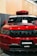 A front view of a customized red SUV with black accents. The car has a prominent grille with multiple horizontal slots, a hood scoop, and a roof-mounted cargo box. The windshield has decals, including 'STNCSG,' and a piece of paper placed on the dashboard. The background includes a wall with vertical wooden panels and part of a ceiling structure.
