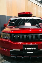 A front view of a customized red SUV with black accents. The car has a prominent grille with multiple horizontal slots, a hood scoop, and a roof-mounted cargo box. The windshield has decals, including 'STNCSG,' and a piece of paper placed on the dashboard. The background includes a wall with vertical wooden panels and part of a ceiling structure.