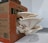 A cluster of large, white oyster mushrooms grows out of an open cardboard box labeled 'Life Cykel'. The mushrooms have wide, curved caps with delicate, pleated gills and are situated on a white paper towel in an indoor setting.
