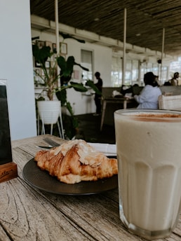 A wooden table holds a plate with a croissant sprinkled with almonds beside a glass filled with a creamy beverage. In the background, a spacious, well-lit cafe features patrons sitting on chairs and a large potted plant against the wall.