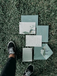 A set of elegant, floral-themed invitations or stationary is laid out on a grassy lawn. The design features peacock illustrations and intricate patterns. A person wearing black pants and black sneakers with white laces is standing next to the invitations.