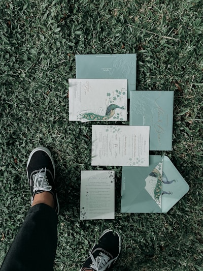 A set of elegant, floral-themed invitations or stationary is laid out on a grassy lawn. The design features peacock illustrations and intricate patterns. A person wearing black pants and black sneakers with white laces is standing next to the invitations.