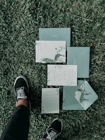 A set of elegant, floral-themed invitations or stationary is laid out on a grassy lawn. The design features peacock illustrations and intricate patterns. A person wearing black pants and black sneakers with white laces is standing next to the invitations.