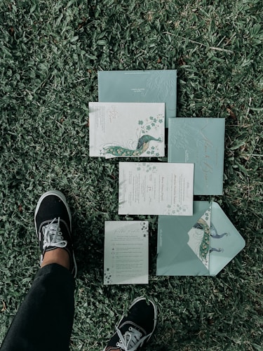 A set of elegant, floral-themed invitations or stationary is laid out on a grassy lawn. The design features peacock illustrations and intricate patterns. A person wearing black pants and black sneakers with white laces is standing next to the invitations.