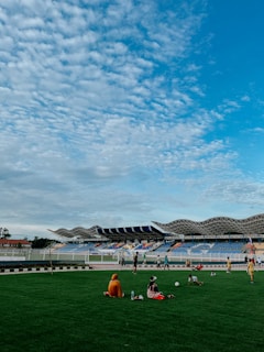 A vibrant open stadium with a modern architectural design featuring a corrugated roof. The lush green field is surrounded by a colorful seating area. Several children are playing or engaged in activities on the grass, with some seated and others standing. The sky is filled with scattered, fluffy clouds under bright blue tones, adding to the lively atmosphere.
