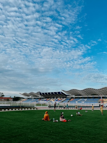 A vibrant open stadium with a modern architectural design featuring a corrugated roof. The lush green field is surrounded by a colorful seating area. Several children are playing or engaged in activities on the grass, with some seated and others standing. The sky is filled with scattered, fluffy clouds under bright blue tones, adding to the lively atmosphere.
