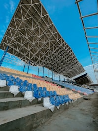 A stadium seating area with multicolored plastic chairs covered in protective plastic wrapping. The seats are arranged in rows with colors blue, red, and beige. Above the seating, there is a large, intricate metal roof structure. The sky is clear and blue, providing a vivid contrast to the metal and the colorful seats.