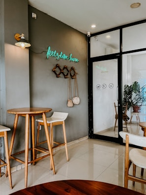 A cozy cafe interior featuring wooden high tables and chairs against a gray wall. A neon sign reading 'feels like home' adds a welcoming touch. Decor includes hanging woven items and a potted plant, reflecting a minimalist aesthetic.