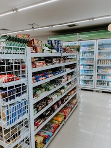 The image depicts the interior of a convenience store with neatly arranged shelves filled with various snacks, packaged foods, and beverages. There are several refrigerated sections visible in the background, stocked with drinks and dairy products. The store appears to be well-lit with a clean and organized layout.