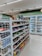 The image depicts the interior of a convenience store with neatly arranged shelves filled with various snacks, packaged foods, and beverages. There are several refrigerated sections visible in the background, stocked with drinks and dairy products. The store appears to be well-lit with a clean and organized layout.