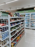 The image depicts the interior of a convenience store with neatly arranged shelves filled with various snacks, packaged foods, and beverages. There are several refrigerated sections visible in the background, stocked with drinks and dairy products. The store appears to be well-lit with a clean and organized layout.