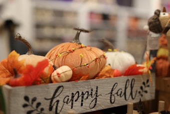 A decorative setting with three pumpkins in a wooden box adorned with the words 'happy fall'. The pumpkins vary in color from orange to white and are surrounded by artificial autumn leaves and small colorful beads on a string.