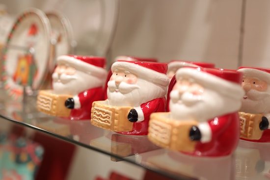 Several ceramic Santa Claus mugs are displayed on a glass shelf. Each mug features Santa holding a decorative block with a cheerful expression. The background includes other blurred festive items.