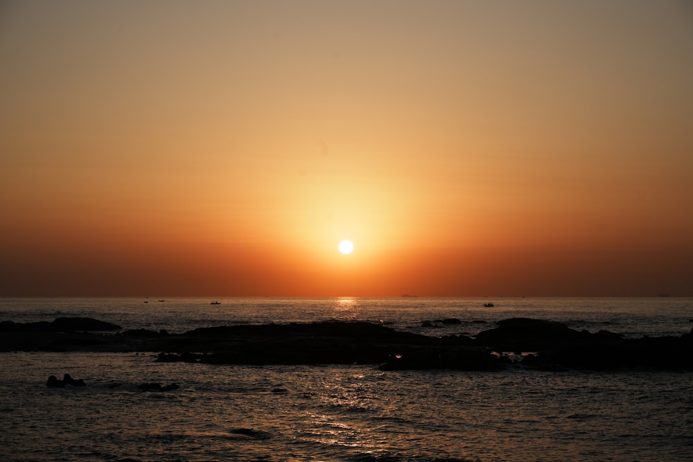 the sun is setting over the ocean with rocks in the foreground