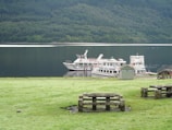 Several cruise boats are docked beside a calm lake with a backdrop of lush green forest-covered hills. The foreground features a wooden picnic table on a grassy area, creating a serene outdoor setting.