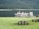Several cruise boats are docked beside a calm lake with a backdrop of lush green forest-covered hills. The foreground features a wooden picnic table on a grassy area, creating a serene outdoor setting.