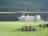 Several cruise boats are docked beside a calm lake with a backdrop of lush green forest-covered hills. The foreground features a wooden picnic table on a grassy area, creating a serene outdoor setting.