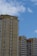 Two multi-story residential buildings with beige and yellow exteriors stand against a backdrop of bright blue sky with scattered clouds. The buildings feature rows of windows, balconies, and architectural details at the roofline.