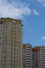 Two multi-story residential buildings with beige and yellow exteriors stand against a backdrop of bright blue sky with scattered clouds. The buildings feature rows of windows, balconies, and architectural details at the roofline.