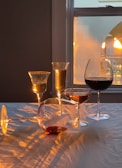 The setting features a variety of wine glasses filled with different types of wine, illuminated by warm, ambient light from a nearby window. The interplay of light and shadow creates patterns on the tablecloth. One glass is tipped over, giving a casual and inviting atmosphere.