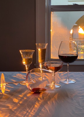 The setting features a variety of wine glasses filled with different types of wine, illuminated by warm, ambient light from a nearby window. The interplay of light and shadow creates patterns on the tablecloth. One glass is tipped over, giving a casual and inviting atmosphere.