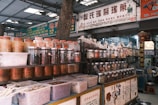 An indoor market scene with numerous jars and containers filled with various dried herbs, spices, and medicinal ingredients. Signs with Chinese characters are displayed above and around the shelves. The area appears organized, with plastic containers on lower shelves and glass jars on higher shelves.
