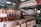 An indoor market scene with numerous jars and containers filled with various dried herbs, spices, and medicinal ingredients. Signs with Chinese characters are displayed above and around the shelves. The area appears organized, with plastic containers on lower shelves and glass jars on higher shelves.