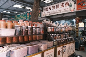 An indoor market scene with numerous jars and containers filled with various dried herbs, spices, and medicinal ingredients. Signs with Chinese characters are displayed above and around the shelves. The area appears organized, with plastic containers on lower shelves and glass jars on higher shelves.