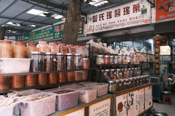 An indoor market scene with numerous jars and containers filled with various dried herbs, spices, and medicinal ingredients. Signs with Chinese characters are displayed above and around the shelves. The area appears organized, with plastic containers on lower shelves and glass jars on higher shelves.