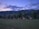 A row of small wooden cabins is situated in a grassy area with a mountainous backdrop. The sky displays a gradient from light to dark, suggesting twilight or early dawn. Trees dot the landscape around the cabins, adding a natural and serene atmosphere.
