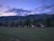 A row of small wooden cabins is situated in a grassy area with a mountainous backdrop. The sky displays a gradient from light to dark, suggesting twilight or early dawn. Trees dot the landscape around the cabins, adding a natural and serene atmosphere.