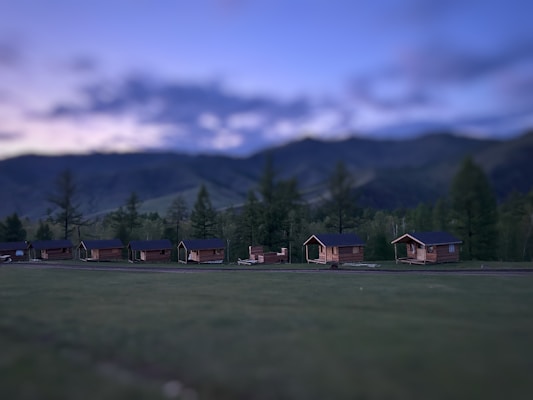 A row of small wooden cabins is situated in a grassy area with a mountainous backdrop. The sky displays a gradient from light to dark, suggesting twilight or early dawn. Trees dot the landscape around the cabins, adding a natural and serene atmosphere.