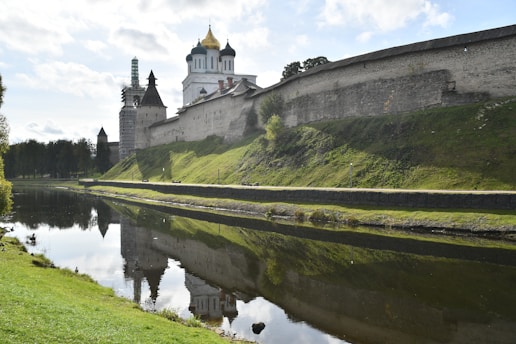 A medieval fortress with tall stone walls runs parallel to a calm body of water, reflecting the structure and the cloudy sky above. A church with golden domes and white towers stands prominently within the fortress walls. The landscape around the fortress is lush with green grass and trees, with the sunlight casting soft shadows and highlighting the textures of the stone and foliage.