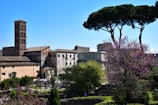 A scenic view of a historic area with ancient Roman architecture, featuring a tall bell tower, several classical buildings, and lush greenery, including blooming trees and distinct tall pine trees. People can be seen in the background, possibly visiting or touring the site.