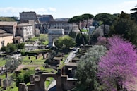 A scenic view of an ancient Roman site with historic ruins and the Colosseum in the background. The landscape includes lush greenery and blooming trees with pink and green hues. Several pathways and older structures are visible, surrounded by vibrant natural elements.