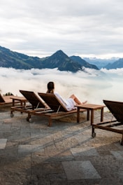 A person in a white bathrobe is reclining on a wooden lounge chair on a stone patio, overlooking a picturesque view of mountains partially covered by clouds. The setting appears calm and serene, with additional empty lounge chairs lined up beside the person.
