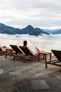 A person in a white bathrobe is reclining on a wooden lounge chair on a stone patio, overlooking a picturesque view of mountains partially covered by clouds. The setting appears calm and serene, with additional empty lounge chairs lined up beside the person.