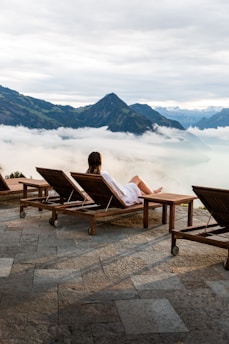 A person in a white bathrobe is reclining on a wooden lounge chair on a stone patio, overlooking a picturesque view of mountains partially covered by clouds. The setting appears calm and serene, with additional empty lounge chairs lined up beside the person.