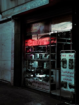 A shopfront featuring a variety of camera equipment, with a prominent red neon 'Canon' sign. The dimly lit display includes cameras, lenses, and related accessories. Signage in a foreign script is visible both above the shop and on a standing board outside.