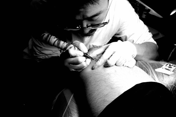 A person wearing glasses and a white shirt is intensely focused on creating a tattoo on another person's arm. The scene is well-lit, emphasizing the concentration and precision required in tattoo artistry. Latex gloves and a tattoo machine are visible, highlighting the hygienic practices in place.