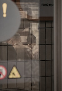 A scene viewed through a window, with reflections and silhouettes of people engaging in conversation. The window is lined with tiles and various signs and symbols are visible on the glass, including an exclamation mark and a caution sign. The atmosphere is muted and somewhat mysterious.