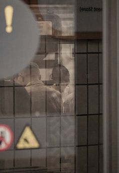 A scene viewed through a window, with reflections and silhouettes of people engaging in conversation. The window is lined with tiles and various signs and symbols are visible on the glass, including an exclamation mark and a caution sign. The atmosphere is muted and somewhat mysterious.